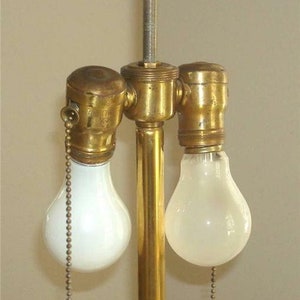 May include: A brass lamp base with two light bulb sockets and a decorative finial on top. The lamp base has a gold finish and the light bulbs are white.
