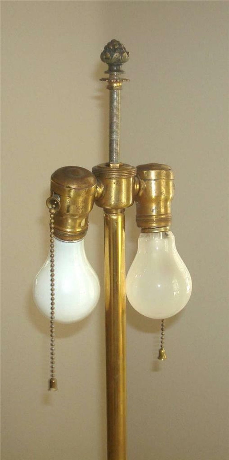 May include: A brass lamp base with two light bulb sockets and a decorative finial on top. The lamp base has a gold finish and the light bulbs are white.