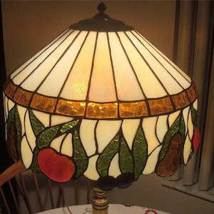 May include: A stained glass lampshade with a white background and a design of green leaves, red and orange fruit, and brown pears. The shade has a gold trim around the top.