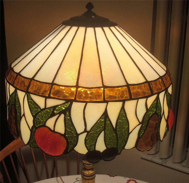 May include: A stained glass lampshade with a white background and a design of green leaves, red and orange fruit, and brown pears. The shade has a gold trim around the top.