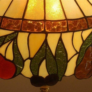 May include: A stained glass lampshade with a floral design featuring red cherries, green leaves, and brown pears. The shade is illuminated from within, casting a warm glow.