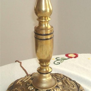 May include: A gold table lamp with a decorative base featuring a floral design. The base is made of metal and has a black finish. The lamp has a black cord.