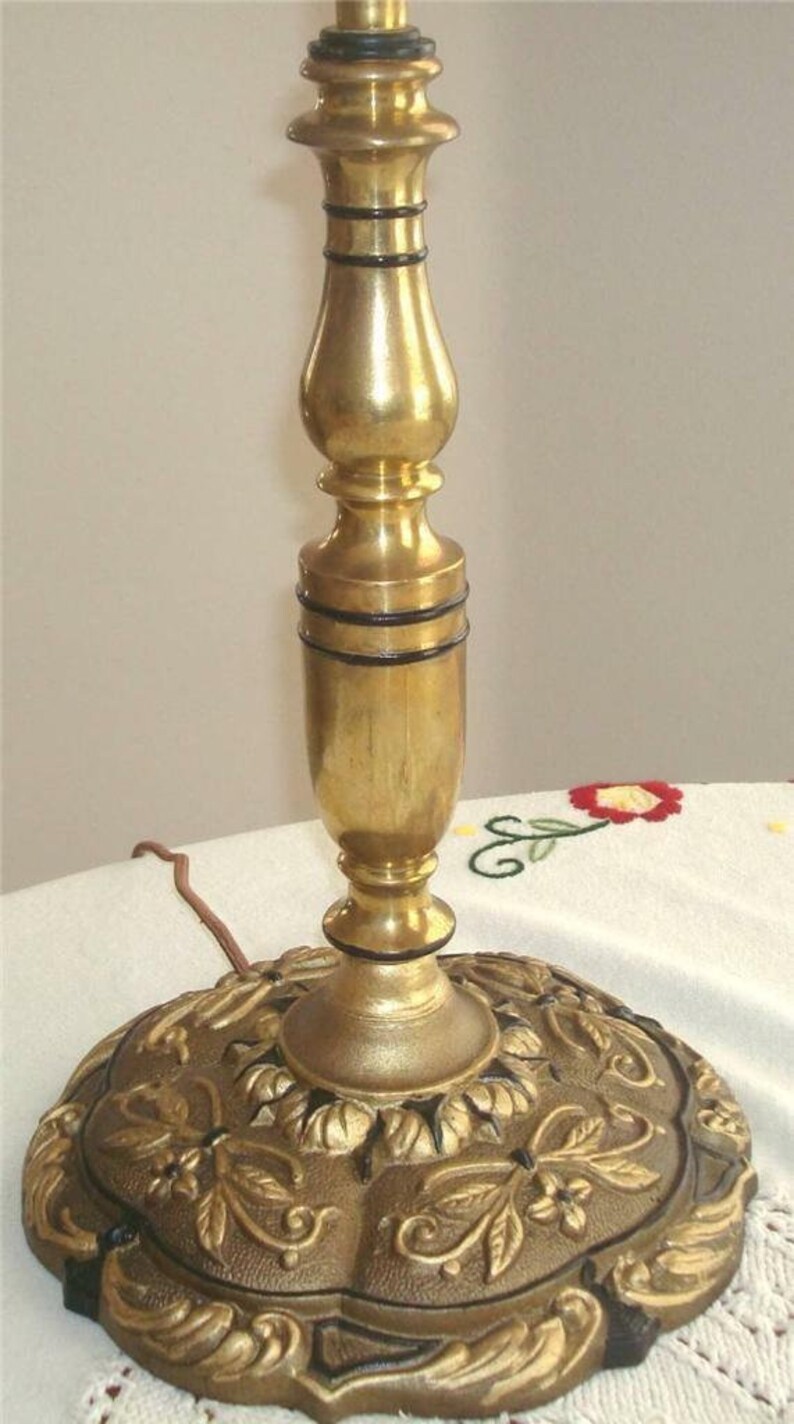 May include: A gold table lamp with a decorative base featuring a floral design. The base is made of metal and has a black finish. The lamp has a black cord.