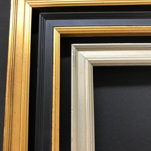 May include: Three picture frames, one gold, one black, and one silver, are stacked on top of each other. The frames are all made of wood and have a simple design.