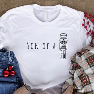May include: White t-shirt with the text "SON OF A" and a black and white illustration of a nutcracker.