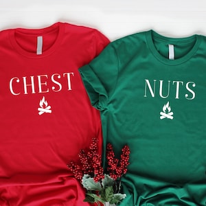 May include: Two t-shirts with a Christmas-themed design. The red shirt says "CHEST" with a campfire graphic. The green shirt says "NUTS" with a campfire graphic.