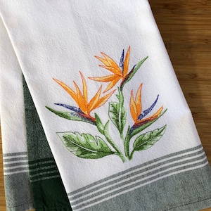 May include: White kitchen towel with an embroidered Bird of Paradise flower design. The flowers are orange and blue with green leaves. The towel has a green and white striped border.