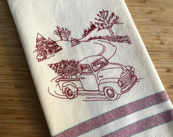 Puede incluir: Un diseño bordado en rojo de una camioneta vintage que lleva un árbol de Navidad, conduciendo por un camino nevado con una granja en el fondo.