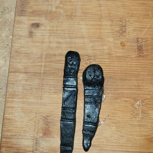 Peut inclure: Deux objets noirs texturés, possiblement faits à la main, sont présentés sur une surface en bois. Les objets ont une forme rectangulaire avec un sommet arrondi et un fond pointu. La surface présente un veinage naturel.