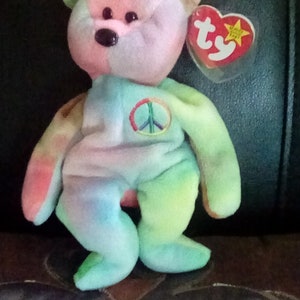 Puede incluir: Un oso de peluche tie-dye con un símbolo de paz en el pecho. El oso es rosa, verde, azul y amarillo. Tiene una etiqueta Ty roja con una estrella blanca.