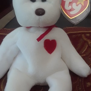 Può includere: Orso di peluche bianco con un cuore rosso sul petto e un nastro rosso intorno al collo. L'orso ha un'etichetta Ty attaccata all'orecchio che dice "Beanie Baby".