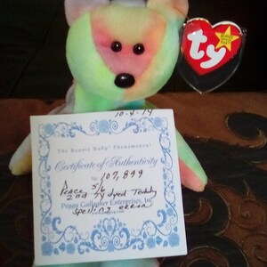 May include: A tie-dye plush toy bear with a certificate of authenticity. The certificate is titled "The Beanie Baby Phenomenon!" and has the number 107,899. The certificate also states "Pencha 5/4 dyed Teddy 2nd" and "Peggy Gallagher Enterprises, Inc. Spelling ekon".