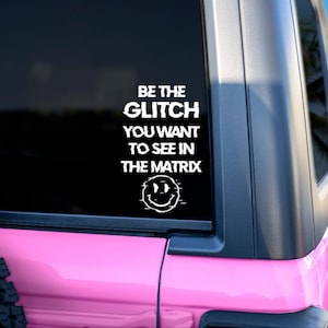 May include: A white vinyl decal on a pink vehicle window. The decal reads "BE THE GLITCH YOU WANT TO SEE IN THE MATRIX" with a glitch-style smiley face. The vehicle has a black trim and a black tire.