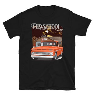 Puede incluir: Una camiseta negra con el texto "Old School" impreso en plata en la parte delantera. La camiseta presenta una camioneta naranja vintage con una cabina blanca y una cama de madera. La camioneta está estacionada frente a un pequeño edificio con un techo rojo.