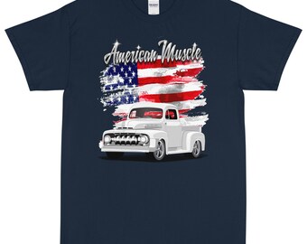 Könnte beinhalten: Ein marineblaues T-Shirt mit einem weißen Vintage-Pickup vor einem verwitterten amerikanischen Flaggen-Motiv. Der Text "American Muscle" befindet sich über dem Truck.