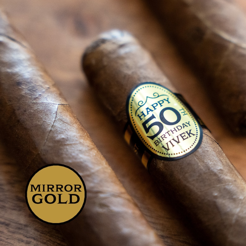 K&ouml;nnte beinhalten: Nahaufnahme von drei braunen Zigarren. Eine Zigarre hat ein gold-schwarzes Etikett mit der Aufschrift "HAPPY 50TH BIRTHDAY VIVEK". Ein goldener Kreis mit den Worten "MIRROR GOLD" befindet sich in der linken unteren Ecke. Die Zigarren liegen auf einer Holzoberfl&auml;che.