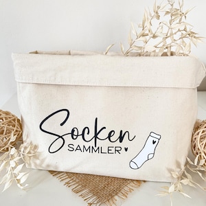 Könnte beinhalten: Eine beige Canvas-Aufbewahrungstasche mit den Worten "Socken SAMMLER" in schwarzer Schrift und einer kleinen Sockenillustration. Die Tasche ist rechteckig mit umgeklapptem Oberteil und mit getrockneten Pflanzen dekoriert.