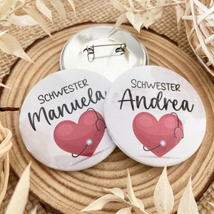 Könnte beinhalten: Zwei weiße runde Knöpfe mit roten Herzen und einem Stethoskop. Der Text auf den Knöpfen lautet "Schwester Manuela" und "Schwester Andrea".