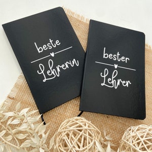 Könnte beinhalten: Zwei schwarze Notizbücher mit weißem Text. Auf einem Notizbuch steht "beste Lehrerin" und auf dem anderen "bester Lehrer". Beide Notizbücher haben ein Herzsymbol über dem Text. Die Notizbücher liegen auf einer beigen Oberfläche.