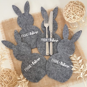 Könnte beinhalten: Graue Hasen-Tischsets aus Filz mit dem weißen Schriftzug "FROHE OSTERN". Auf einem der Tischsets liegen eine Gabel und ein Messer. Die Tischsets liegen auf einer Jutefläche, mit einem dekorativen Garnball und Trockenblumen.