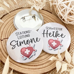 Könnte beinhalten: Zwei weiße runde Knöpfe mit roten Herzen und medizinischen Symbolen. Ein Knopf sagt "Stations-Leitung Simone" und der andere sagt "Schwester Heike".
