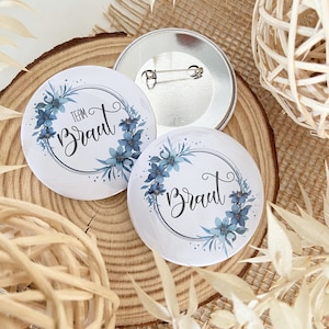 Könnte beinhalten: Zwei weiße runde Knöpfe mit blauen Blumenmustern und dem Text "Team Braut" auf einem Knopf und "Braut" auf dem anderen. Die Knöpfe haben eine silberne Nadel auf der Rückseite.