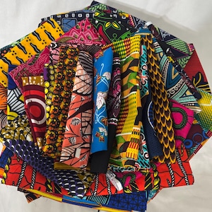 May include: A collection of colorful African wax print fabrics, fanned out to display the variety of patterns. The fabrics feature bold geometric and floral designs in vibrant hues of red, yellow, blue, and green. The textiles are folded and arranged to showcase the diverse patterns.