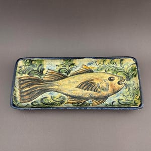 May include: A rectangular ceramic tray featuring a hand-painted golden-yellow fish centered against a background of green and blue swirling seaweed patterns. The tray has a dark blue rim and a textured, mottled finish across the entire surface.