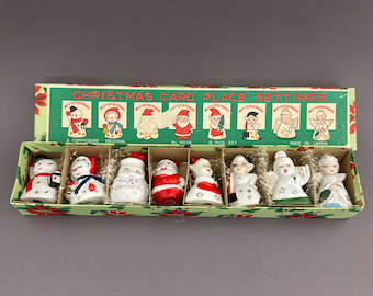 Może przedstawiać: Zestaw ośmiu ceramicznych wizytówek świątecznych w zielonym pudełku z napisem CHRISTMAS CARD PLACE SETTINGS. Zestaw zawiera miniaturowe figurki bałwanów, Świętego Mikołaja i aniołów, każda z malowanymi detalami i w świątecznych kolorach.