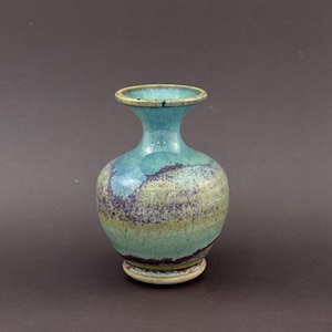 May include: A small ceramic vase with a flared rim and a bulbous body. The glaze features a blend of turquoise, light green, and deep purple hues, creating a mottled, layered appearance across the surface of the vessel with a smooth finish.