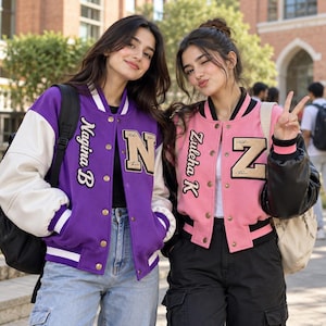 Pode incluir: Uma jaqueta universitária roxa com mangas brancas, o texto Nagina B e um patch com a letra N, ao lado de uma jaqueta rosa com mangas pretas, o texto Zuleha K e um patch com a letra Z. Ambas as jaquetas possuem botões de pressão e punhos canelados.