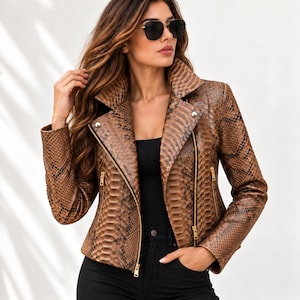 May include: A brown and black snakeskin-patterned biker jacket featuring a wide notched collar, long sleeves, and gold-toned metal zips on the front closure and side pockets. The jacket is worn over a simple black top and black trousers.