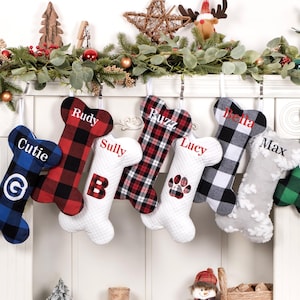 May include: Seven personalized dog bone-shaped Christmas stockings with different plaid patterns, including red and black, black and white, and green and black. The stockings are hung on a white fireplace mantel and are labeled with the names "Cutie", "Rudy", "Sully", "Buzz", "Lucy", "Bella", and "Max".