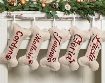 May include: Five bone-shaped stockings made of light beige fabric hanging from a branch. Each stocking features a red and black buffalo plaid name printed in cursive: Cordyn, Mallorie, Jordan, Gloria, and Lucia, with paw print accents.