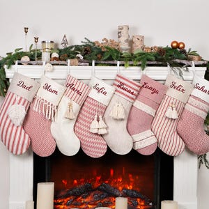 May include: Eight personalized Christmas stockings hang above a fireplace. Each stocking is a different pattern and color, with names embroidered in a matching thread. The stockings are decorated with tassels and other embellishments. The fireplace has a faux fire and candles.