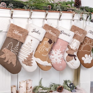 May include: A set of six Christmas stockings in various colors, including brown, white, and pink, each personalized with a name. The stockings feature festive designs like a bear, Christmas tree, snowman, and snowflake, with faux fur cuffs.