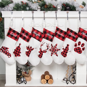 May include: Set of eight white quilted Christmas stockings with red and black buffalo plaid trim. Each stocking is personalized with a name and features a different festive design, including a Christmas tree, angel, reindeer, snowflake, snowman, paw print, and a truck.
