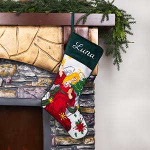 K&ouml;nnte beinhalten: Ein Weihnachtsstrumpf mit dem Namen "Luna" auf einem dunkelgr&uuml;nen Samtoberteil gestickt. Der Strumpf zeigt ein detailliertes Gobelindesign eines Engels in einem roten Kleid mit goldenen Akzenten und wei&szlig;en Fl&uuml;geln auf wei&szlig;em Hintergrund.