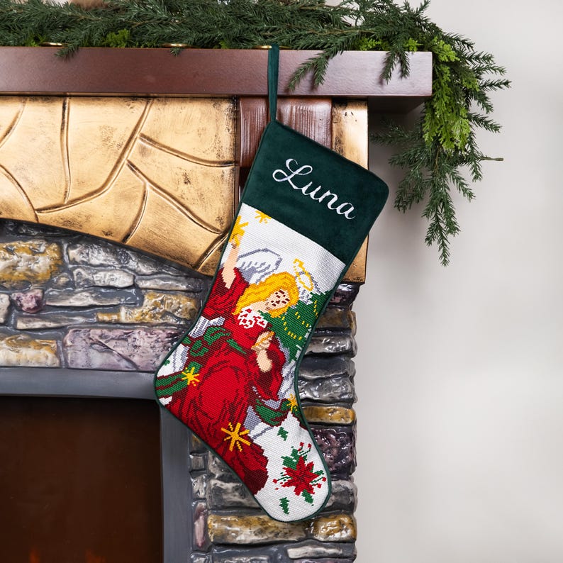 K&ouml;nnte beinhalten: Ein Weihnachtsstrumpf mit dem Namen "Luna" auf einem dunkelgr&uuml;nen Samtoberteil gestickt. Der Strumpf zeigt ein detailliertes Gobelindesign eines Engels in einem roten Kleid mit goldenen Akzenten und wei&szlig;en Fl&uuml;geln auf wei&szlig;em Hintergrund.