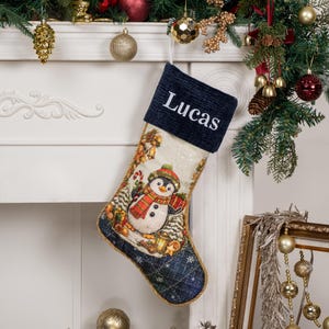 Op de afbeelding: Een kerstkous met een donkerblauwe ribfluwelen boord waarop de naam Lucas in het wit is geborduurd. Het lichaam van de kous toont een geweven patroon van een pingu&iuml;n met een muts en sjaal, omringd door kerstversieringen.