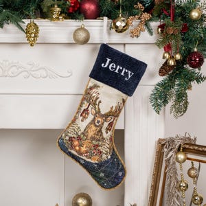 Op de afbeelding: Een kerstkous met een marineblauwe ribfluwelen boord waarop de naam Jerry in het wit is geborduurd. Het lichaam toont een geweven wandtapijt van een hert omringd door dennenappels en groen, afgewerkt met een gouden gevlochten rand.
