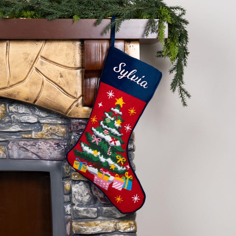 K&ouml;nnte beinhalten: Ein roter Weihnachtsstrumpf mit marineblauem B&uuml;ndchen, personalisiert mit dem Namen "Sylvia" in wei&szlig;er Schrift. Der Strumpf zeigt ein detailliertes gesticktes Weihnachtsbaumdesign mit Geschenken, Sternen und Schneeflocken. Eine festliche Weihnachtsdekoration.