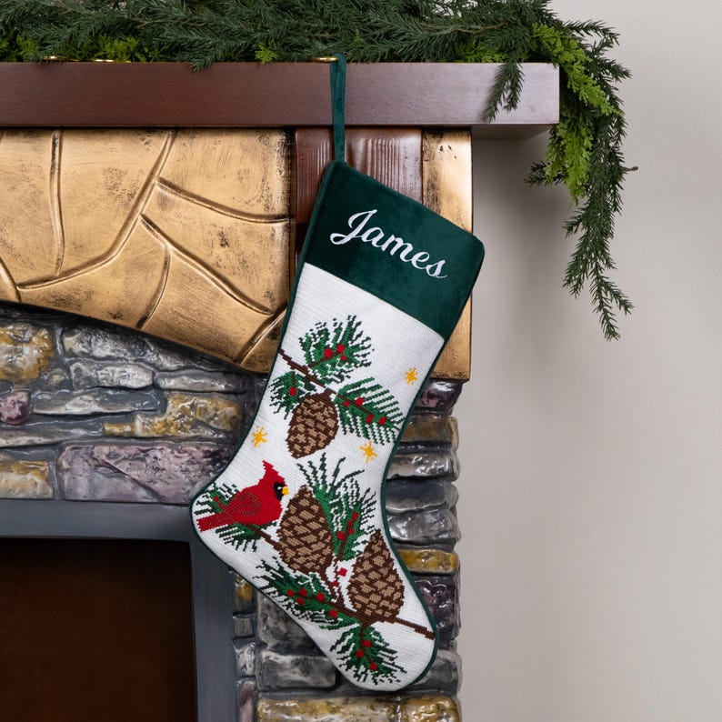 K&ouml;nnte beinhalten: Ein Weihnachtsstrumpf mit einem gr&uuml;nen Samt-Oberteil und dem Namen "James" in Wei&szlig; gestickt. Der wei&szlig;e K&ouml;rper zeigt ein detailliertes Nadelspitzendesign aus Tannenzapfen, einem roten Kardinal und immergr&uuml;nen Zweigen.