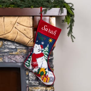 K&ouml;nnte beinhalten: Ein Weihnachtsstrumpf mit rotem Samt-Oberteil, bestickt mit dem Namen "Sarah". Der Strumpf zeigt einen Eisb&auml;ren mit rotem Schal und Hut, umgeben von einer verschneiten Landschaft, Weihnachtsb&auml;umen und Geschenken. Der Hintergrund ist marineblau.