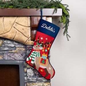 K&ouml;nnte beinhalten: Ein Weihnachtsstrumpf mit Rentier-Design. Der Strumpf ist rot mit einer blauen Manschette, auf der der Name "Debbie" gestickt ist. Das Rentier hat einen gr&uuml;n-wei&szlig;en Schal und tr&auml;gt ein Geschenk. Er h&auml;ngt an einem Kamin.
