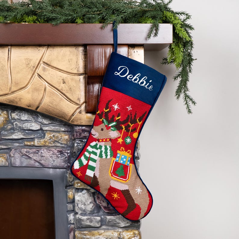 K&ouml;nnte beinhalten: Ein Weihnachtsstrumpf mit Rentier-Design. Der Strumpf ist rot mit einer blauen Manschette, auf der der Name "Debbie" gestickt ist. Das Rentier hat einen gr&uuml;n-wei&szlig;en Schal und tr&auml;gt ein Geschenk. Er h&auml;ngt an einem Kamin.