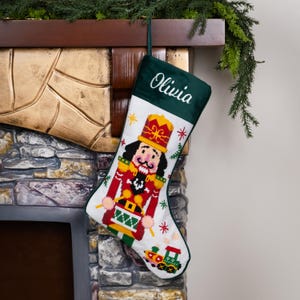 K&ouml;nnte beinhalten: Ein Weihnachtsstrumpf mit einem gr&uuml;nen Oberteil, auf dem der Name "Olivia" in wei&szlig;er Schrift gestickt ist. Der Hauptteil zeigt ein Kreuzstichmuster eines Nussknackersoldaten in Rot, Gelb und Gr&uuml;n, mit einem Spielzeugzug am unteren Ende.