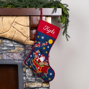 K&ouml;nnte beinhalten: Ein Weihnachtsstrumpf mit roter Manschette und dem Namen "Kyle" in Wei&szlig; gestickt. Der Strumpf ist marineblau und mit einem festlichen Design mit dem Weihnachtsmann in seinem Schlitten, Rentieren, Schneeflocken und einem gelben Mond verziert.