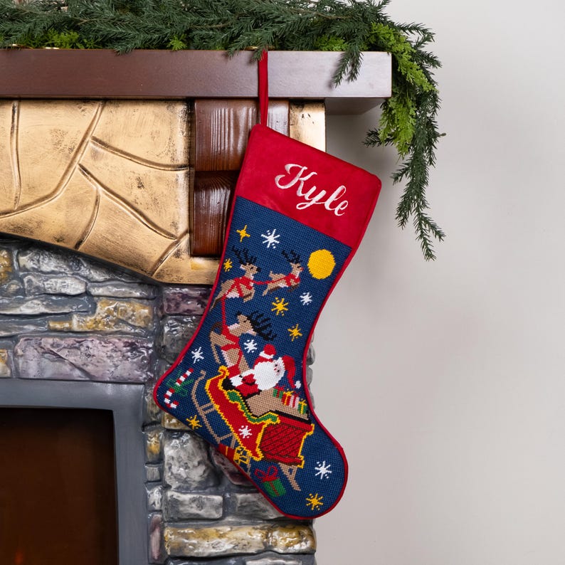 K&ouml;nnte beinhalten: Ein Weihnachtsstrumpf mit roter Manschette und dem Namen "Kyle" in Wei&szlig; gestickt. Der Strumpf ist marineblau und mit einem festlichen Design mit dem Weihnachtsmann in seinem Schlitten, Rentieren, Schneeflocken und einem gelben Mond verziert.