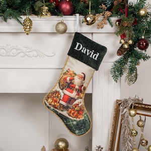 Op de afbeelding: Een kerstkous met een donkergroene ribfluwelen boord waarop de naam David in het wit is geborduurd. Het lichaam van de kous toont een gedetailleerde illustratie van de Kerstman met een zak vol cadeaus, afgewerkt met een gouden koordrand.